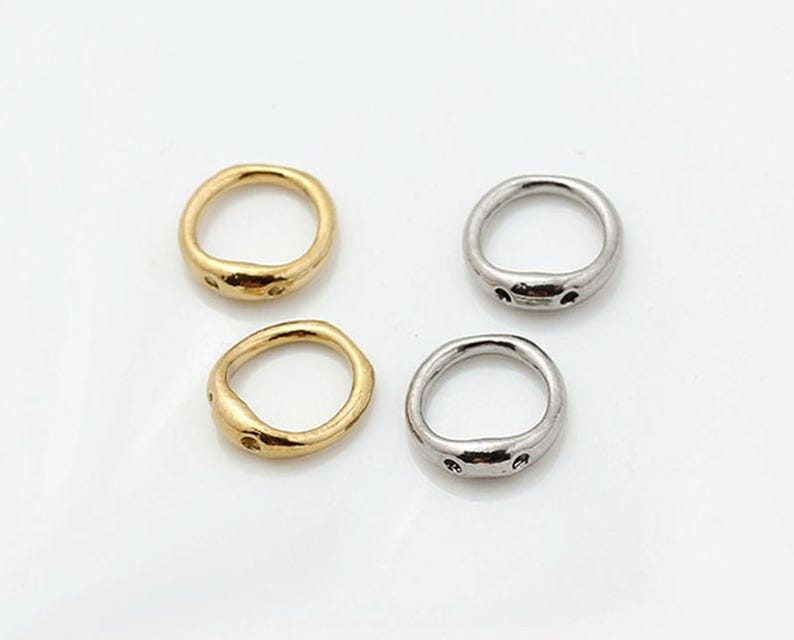 May include: Two gold-toned and two silver-toned metal rings with an irregular, organic shape. Each ring features a slightly flattened section on one side with two small, circular indentations or holes, creating a distinct, textured appearance.