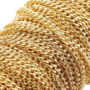 Può includere: Primo piano di un rotolo di catena dorata lucida. Le maglie della catena sono strettamente intrecciate, creando una superficie strutturata. La catena viene probabilmente utilizzata per la creazione di gioielli o altri progetti artigianali.