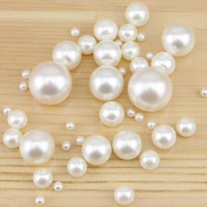 May include: A collection of white, round pearls of varying sizes scattered on a light brown wooden surface. The pearls have a glossy, reflective finish, creating a luminous effect. The arrangement includes both large and small pearls.