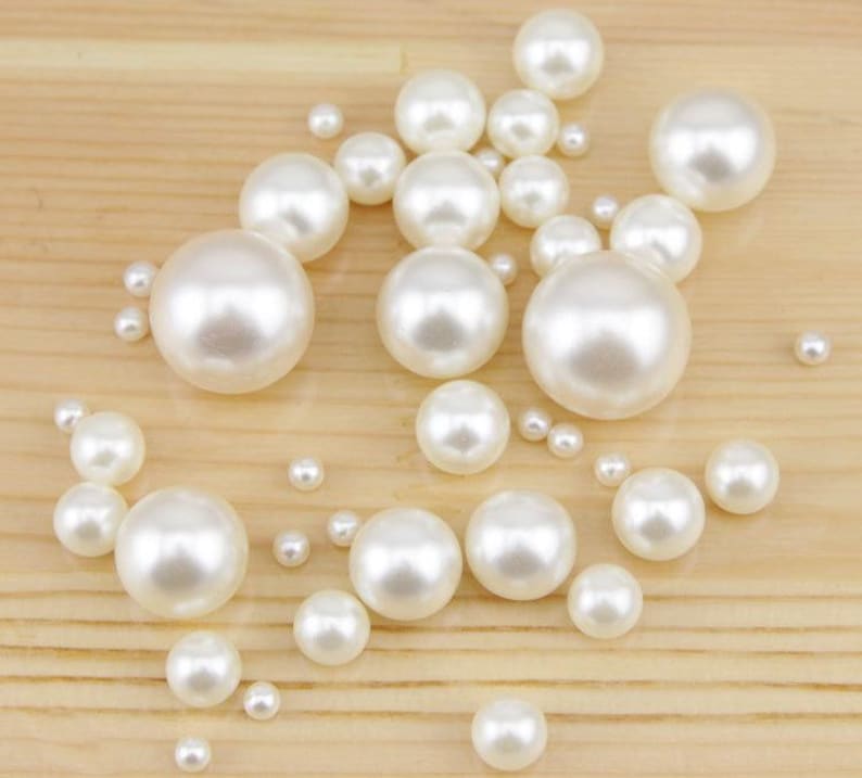 May include: A collection of white, round pearls of varying sizes scattered on a light brown wooden surface. The pearls have a glossy, reflective finish, creating a luminous effect. The arrangement includes both large and small pearls.