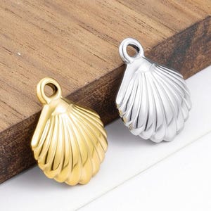 May include: Two seashell-shaped pendants, one gold-toned and one silver-toned, with a loop at the top for a chain. The pendants have a ribbed design and are displayed on a wooden surface.