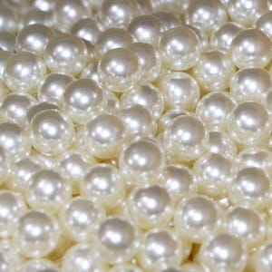 May include: A close-up shot of numerous off-white, round pearls. The pearls have a lustrous, reflective surface, with highlights creating a sense of depth and volume. The pearls are clustered together, filling the frame.
