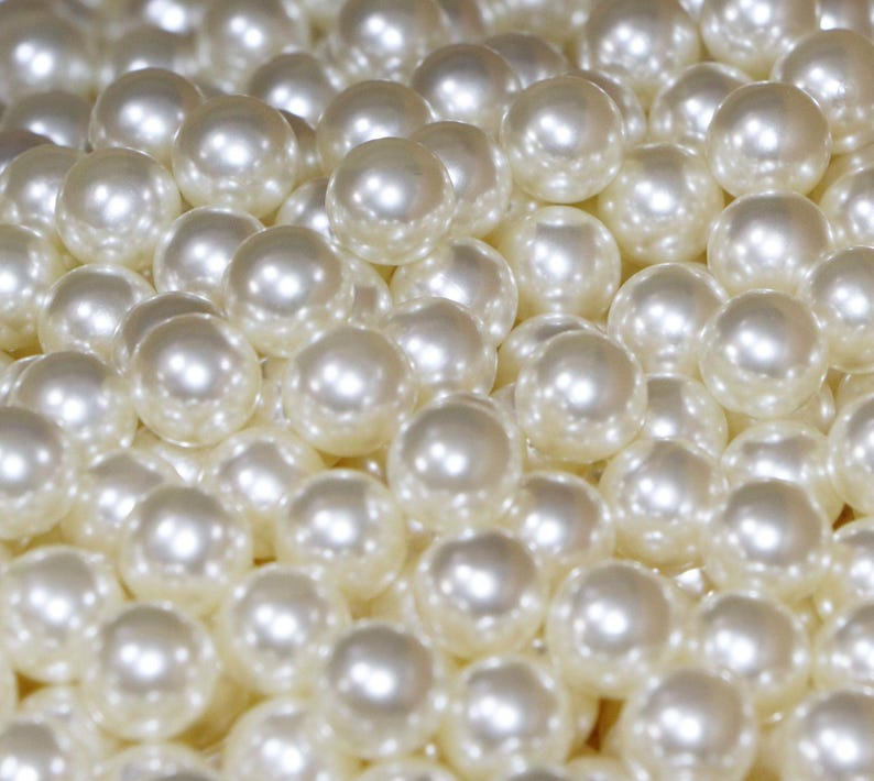 May include: A close-up shot of numerous off-white, round pearls. The pearls have a lustrous, reflective surface, with highlights creating a sense of depth and volume. The pearls are clustered together, filling the frame.