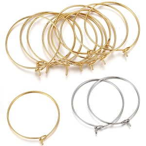 Puede incluir: Una colección de aros metálicos circulares y finos para la creación de joyería. El conjunto incluye varios aros dorados dispuestos en grupo en la parte superior, un aro dorado individual en la parte inferior izquierda y dos aros plateados a la derecha.