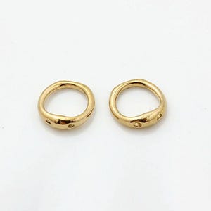 May include: Two small, circular gold-toned metal rings with an irregular, organic shape. Each ring features a smooth, polished surface with two small, evenly spaced circular indentations or holes along the outer edge of the band.