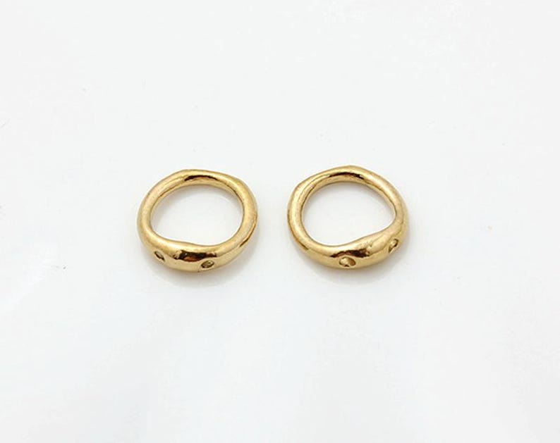 May include: Two small, circular gold-toned metal rings with an irregular, organic shape. Each ring features a smooth, polished surface with two small, evenly spaced circular indentations or holes along the outer edge of the band.
