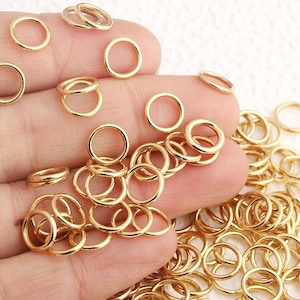 Puede incluir: Una gran colección de pequeñas anillas de salto circulares de metal en tono dorado. Estos fornituras para joyería presentan un acabado liso y pulido con un diseño sencillo de bucle abierto, utilizado para conectar diversos componentes en la creación de joyas.