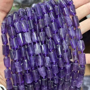 May include: Strand of rectangular amethyst beads in varying shades of purple. The beads are strung together with a matching purple cord. The beads have a polished, translucent appearance, and are held in a hand.