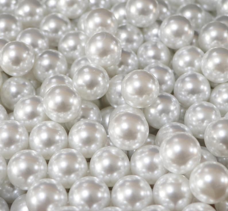 May include: A close-up shot of numerous white, round pearls. The pearls have a lustrous, reflective surface, and are tightly packed together, filling the frame. The pearls are a creamy white color, and the lighting creates highlights and shadows.