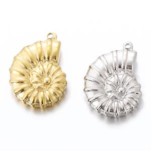 Puede incluir: Dos abalorios de concha de nautilus, uno dorado y otro plateado. Los abalorios son detallados y tienen un diseño en espiral.