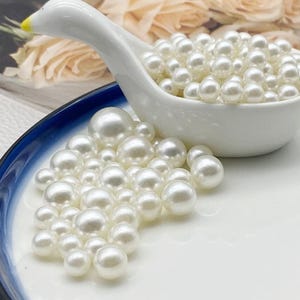 May include: A white ceramic swan-shaped dish filled with numerous white pearls, with additional pearls scattered on a white and blue-rimmed plate. The pearls are round and have a lustrous sheen, suggesting a jewelry or craft theme.