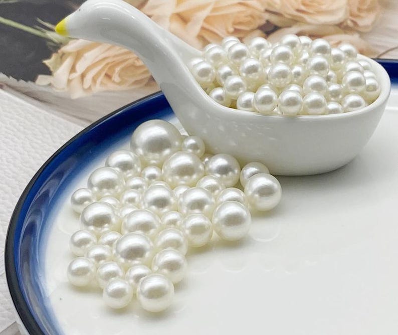 May include: A white ceramic swan-shaped dish filled with numerous white pearls, with additional pearls scattered on a white and blue-rimmed plate. The pearls are round and have a lustrous sheen, suggesting a jewelry or craft theme.