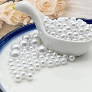 May include: A white ceramic swan-shaped dish filled with numerous white, round pearls. Some pearls are scattered on a white plate with a blue rim. The swan's beak is yellow.