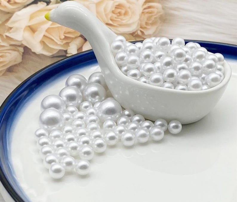 May include: A white ceramic swan-shaped dish filled with numerous white, round pearls. Some pearls are scattered on a white plate with a blue rim. The swan's beak is yellow.