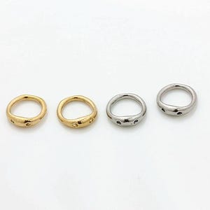 May include: Four rings arranged in a horizontal row. Two rings are gold-toned and two are silver-toned. Each ring features an irregular, organic shape with a smooth, polished surface and two small, circular indentations on the front side.