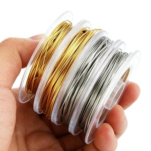 Puede incluir: Cuatro carretes de alambre para manualidades dispuestos en fila, con dos carretes de alambre dorado metálico y dos carretes de alambre plateado metálico. Cada carrete es de plástico transparente y contiene un alambre fino y flexible.