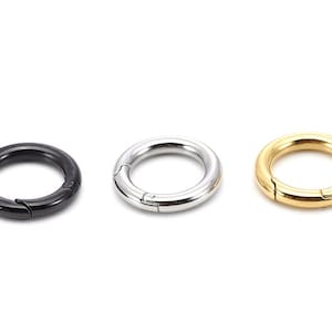 Könnte beinhalten: Drei runde Metall-Federringverschlüsse in Schwarz, Silber und Gold. Jeder Ring hat eine aufklappbare Öffnung zum Befestigen. Die Verschlüsse sind vor einem weißen Hintergrund abgebildet.