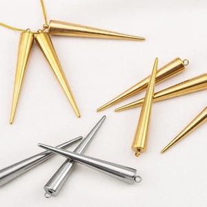 Könnte beinhalten: Eine Sammlung von metallischen spitzen Anhängern in Gold- und Silbertönen. Jeder Anhänger hat eine lange, schlanke, konische Form mit einer glatten, polierten Oberfläche und einer kleinen runden Öse an der Oberseite zur Befestigung an einer Kette.