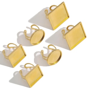 May include: A collection of gold-colored adjustable rings with rectangular and oval settings. The rings are displayed on a white surface, casting shadows. The settings are designed to hold various embellishments.