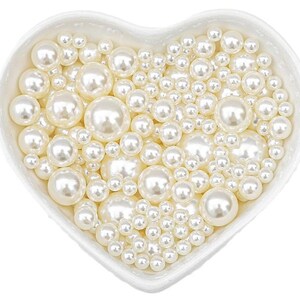 May include: A heart-shaped white ceramic dish filled with various sizes of creamy white faux pearls. The pearls have a lustrous sheen, creating a classic and elegant aesthetic. Ideal for jewelry making or craft projects.