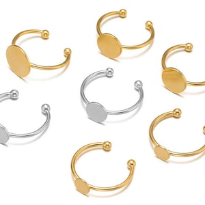 May include: A selection of open-ended rings in gold and silver. Each ring has a small, flat, circular plate and ball-shaped ends. The rings are designed to be adjustable.