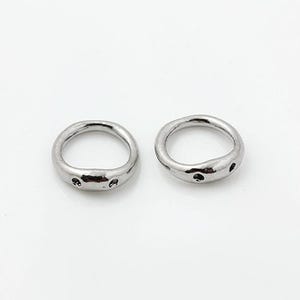 May include: Two silver-toned metal rings with an irregular, organic shape. Each ring features a smooth, polished surface with small, circular indentations or holes along the outer edge, creating a distinct, textured appearance on the band.