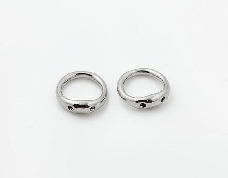 May include: Two silver-toned metal rings with an irregular, organic shape. Each ring features a smooth, polished surface with small, circular indentations or holes along the outer edge, creating a distinct, textured appearance on the band.