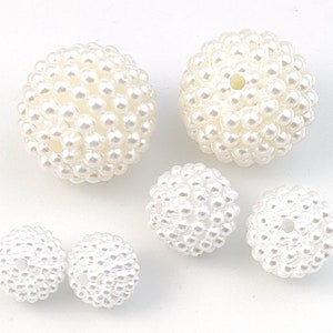 May include: Several white, spherical beads are arranged on a white surface. The beads are covered in smaller, pearl-like embellishments, creating a textured, clustered appearance. Some beads have a hole. These beads are suitable for jewelry making and crafts.