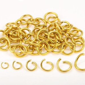 Op de afbeelding: Een verzameling goudkleurige sprongringen in verschillende maten, sommige open en sommige gesloten. De ringen zijn gerangschikt op een witte ondergrond, met een stapel verbonden ringen op de achtergrond en een rij open ringen op de voorgrond.