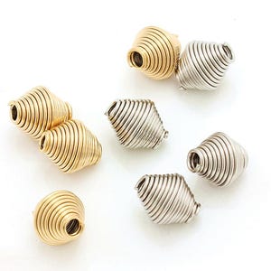 Puede incluir: Cuentas en espiral doradas y plateadas. Las cuentas tienen forma cónica con un agujero central y están hechas de metal enrollado. Se encuentran dispersas sobre una superficie blanca, mostrando su brillo metálico.