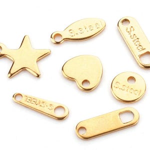 May include: A set of seven gold-colored metal charms with different shapes and sizes. The charms include a star, a heart, a circle, a rectangle, and two oval shapes. Each charm has a different inscription, including "S.Steel", "TEEMUS", and "S.Steel".