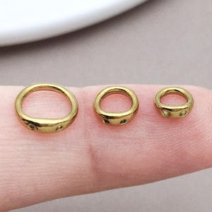 May include: Three gold-toned metal rings of varying sizes arranged in a row. Each ring features a smooth, rounded band with a slightly flattened section at the base containing two small, circular indentations or holes on the front surface.