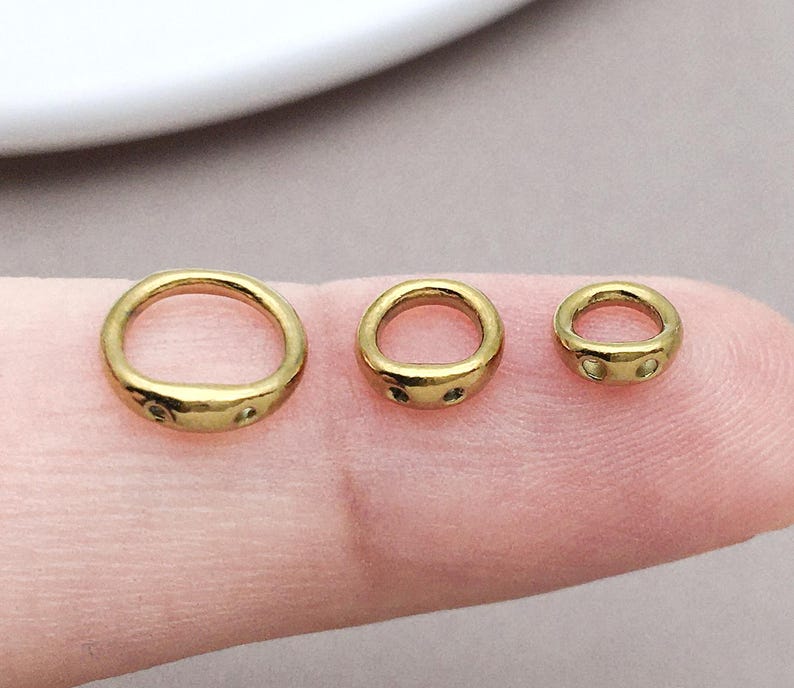 May include: Three gold-toned metal rings of varying sizes arranged in a row. Each ring features a smooth, rounded band with a slightly flattened section at the base containing two small, circular indentations or holes on the front surface.
