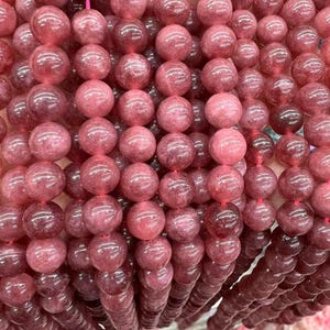 Puede incluir: Primer plano de varios hilos de cuentas redondas y pulidas en tonos rosa y burdeos. Las cuentas están ensartadas en un fino cordón rojo. Las cuentas tienen un acabado liso y brillante y son de tamaño uniforme. Adecuado para la fabricación de joyas.