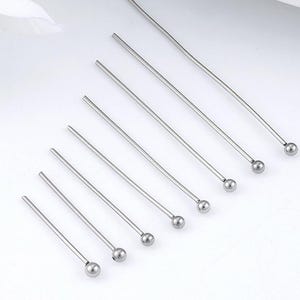 Könnte beinhalten: Eine Sammlung silberfarbener Kopfstifte, jeder mit einer kleinen, runden Kugel an einem Ende. Die Stifte sind diagonal auf einer weißen Oberfläche angeordnet und zeigen ihr schlichtes, elegantes Design. Ideal für die Schmuckherstellung und das Basteln.