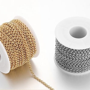 May include: Two spools of chain, one gold and one silver, are wound around white spools. The chain is a small, round link chain.