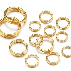 May include: A collection of gold-coloured metal split rings in various sizes. These circular connectors feature a double-loop design, commonly used for keychains or jewellery making, arranged in a scattered pattern against a plain white background.