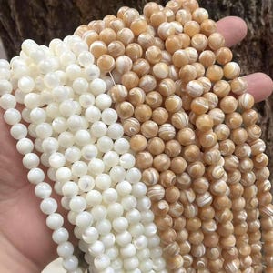 Puede incluir: Primer plano de hileras de cuentas redondas en dos colores: blanco crema y marrón claro. Las cuentas tienen un brillo nacarado y están ensartadas, probablemente para la fabricación de joyas. Las cuentas se sostienen en una mano, con un fondo de madera natural.
