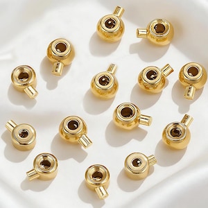 May include: A collection of thirteen small, shiny gold-toned metal beads, each featuring a spherical body with a single protruding cylindrical loop. The beads have a smooth, reflective surface and a central hole, arranged in a scattered pattern.