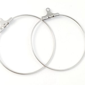 Peut inclure: Deux apprêts de boucles d'oreilles à cerceau argentés. Chaque apprêt a un cerceau circulaire avec un fermoir à charnière en haut. Les cerceaux sont en métal fin et brillant, sur fond blanc.