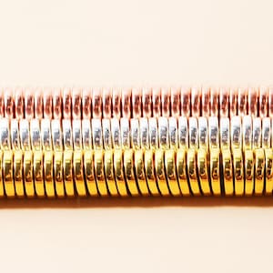 May include: Close-up of three rows of metal coils in copper, silver, and gold colors. The coils are tightly wound and create a textured, metallic surface. The background is a soft, neutral color.