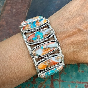 Op de afbeelding: Een zilveren armband met een touwontwerp rand. De armband is versierd met zes ovale stenen van turquoise, koraal en parelmoer.