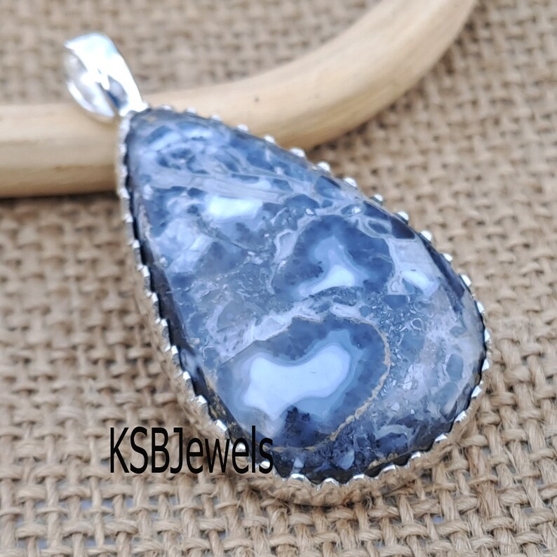 May include: A silver pendant with a blue and white agate stone. The stone has a swirling pattern and is set in a silver bezel. The pendant is on a burlap background. KSBJewels is printed on the bottom of the image.