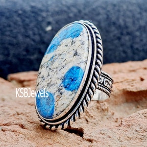 May include: A silver ring featuring a blue and white oval stone set in a rope-like band. The band is decorated with a floral design. KSBJewels is printed on the ring.