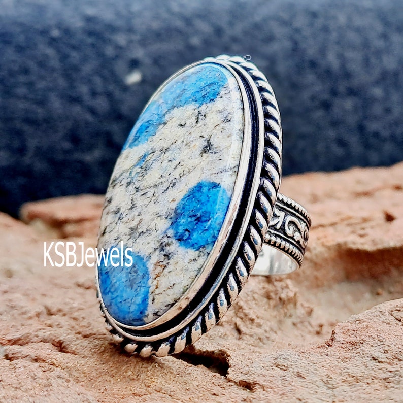 May include: A silver ring featuring a blue and white oval stone set in a rope-like band. The band is decorated with a floral design. KSBJewels is printed on the ring.