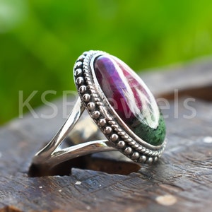 Peut inclure: Une bague en argent avec une pierre ovale verte et rouge sertie dans une bélière à perles. La bague a une monture fendue.