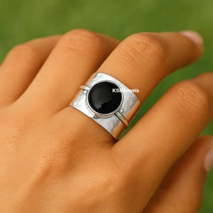 May include: A silver ring with a large, round black stone as the focal point. The band has a hammered texture and is wide, with a smaller band encircling the black stone. The text "KS Jewels" is visible on the ring.