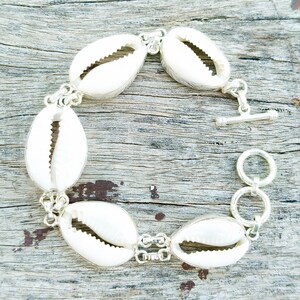May include: A bracelet made of white cowrie shells connected by silver-coloured metal links. The bracelet has a toggle clasp. The shells are arranged in a circular design. The bracelet is displayed on a wooden surface. The bracelet's design is natural.