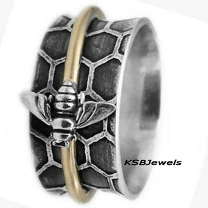 May include: A silver spinning ring with a bee design and a honeycomb pattern. The ring has a gold band that spins around the bee and honeycomb.
