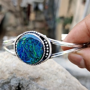 May include: A silver bangle bracelet with a round, green and blue opal cabochon set in a silver bezel. The bezel is decorated with small silver beads.